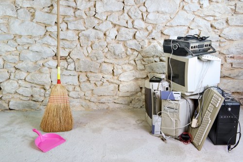 The image shows a cluttered corner of an indoor or storage space with a stone brick wall in the background. On the left side, there is a traditional straw broom with a wooden handle standing upright, and a small pink dustpan resting on the light-colored concrete floor. On the right side, there is an assortment of discarded electronic waste, including an old CRT television, a desktop computer tower, a laptop, a wired keyboard, a small printer, and various cables draped over the items. A large black speaker or amplifier is also visible among the equipment. The scene appears to depict disposal or storage of obsolete electronic devices and cleaning equipment, aligning with rubbish removal services offered by Commercial Waste Removal Putney. The environment suggests a basement, garage, or storage room in a residential or commercial property in Putney or nearby areas, with natural lighting illuminating the scene.