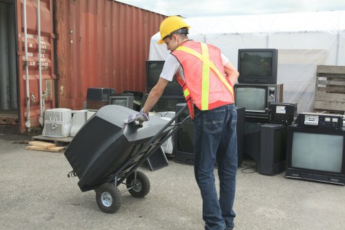 A waste collection worker wearing a yellow safety helmet, a pink high-visibility vest with reflective yellow stripes, a white t-shirt, and blue jeans, is pushing a large black wheeled rubbish bin on a metal frame across an outdoor area. The background features a red shipping container on the left, with various old electronic appliances such as televisions, monitors, and stereo systems stacked against it and on the ground, indicating a disposal or recycling site. The setting appears to be a commercial or industrial waste removal location, possibly in Putney or nearby, associated with the services of Commercial Waste Removal Putney. The scene is lit with natural daylight, emphasizing the textures of the bin, the worker's clothing, and the surrounding objects, all situated on an asphalt surface with some wooden pallets in the background.