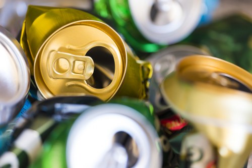Close-up photograph of several crushed and partially flattened aluminium and tin cans in various shades of metallic silver, gold, green, and blue, stacked haphazardly on a surface. The cans exhibit dents, creases, and glossy finishes, with some displaying pull-tab openings and labels that are partially obscured. The background includes blurred out elements suggesting an outdoor environment, such as a pavement or driveway, possibly associated with waste collection or rubbish removal activities in Putney. The image emphasizes the importance of waste sorting and recycling in commercial or domestic waste management, as handled by services like Commercial Waste Removal Putney. The focus on the cans highlights common types of recyclable aluminium and tin waste encountered in rubbish removal services in London suburbs.