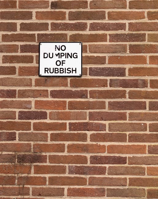 A rectangular white metal sign with black text reading 'NO DUMPING OF RUBBISH' is mounted on a brick wall composed of reddish-brown bricks with darker mortar joints. The bricks have a textured surface
